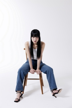 一位 20 多岁的韩国女性，穿着蓝色牛仔裤，坐在椅子上摆姿势，背景为白色 