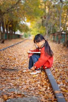 亚洲小女孩深秋季节坐在落叶满地的户外地上看书