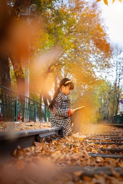 亚洲小女孩深秋季节坐在落叶满地的户外地上看书